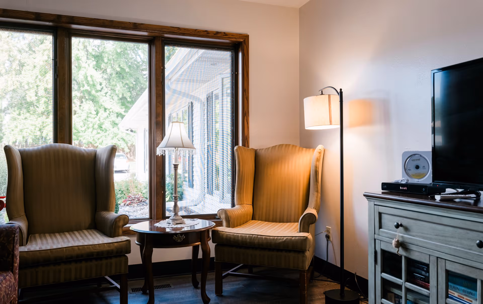 A cozy sitting area with two beige wingback chairs separated by a small round wooden table with a decorative lamp on it. Behind the chairs is a large window showing greenery outside. To the right, there is a floor lamp and a wooden cabinet with a television on top.