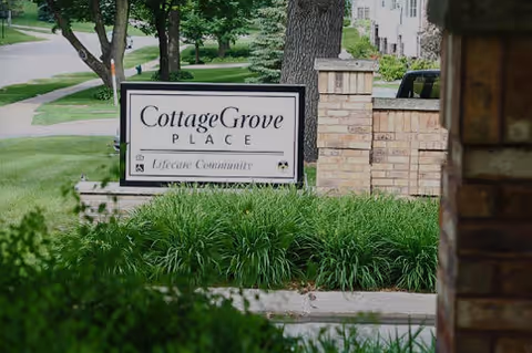 Outdoor view of a sign for Cottage Grove Place Lifecare Community surrounded by green bushes and trees, with a sidewalk and street in the background.