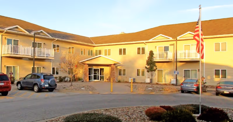 Exterior view of a two-story senior living facility building with beige siding and a brown roof. The entrance has a small covered porch supported by brick columns. Several cars are parked in front of the building, and an American flag is displayed on a flagpole near the entrance. There are small trees and landscaping with rocks and bushes around the parking area.
