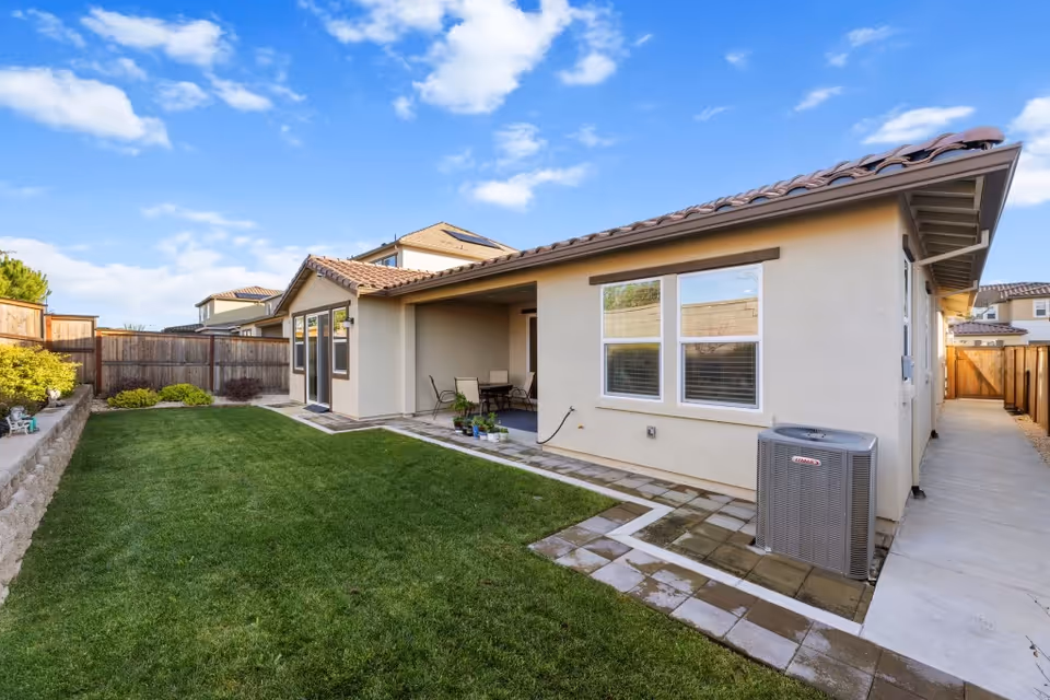 Backyard of a single-story house with a grassy lawn, paved patio seating area, and an outdoor air conditioning unit.