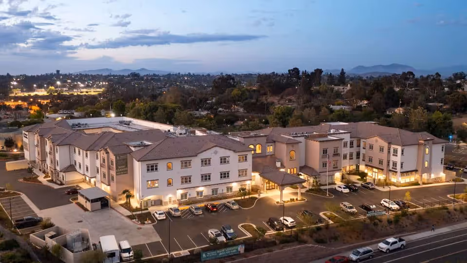 Aerial dusk view of a large illuminated senior living building with parking lot and surrounding neighborhood.