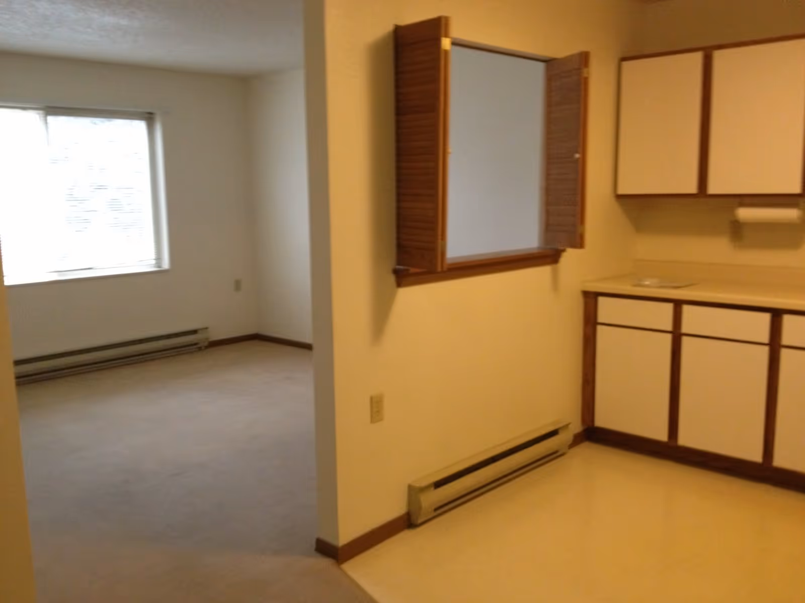 Interior view of an apartment showing a small kitchen area with white cabinets and wooden trim on the right, and an adjacent empty room with a window and beige carpet on the left.