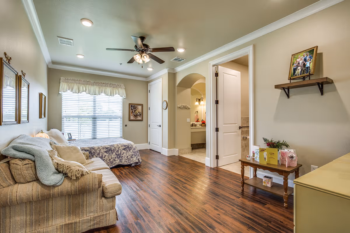 A cozy bedroom in Avalon Memory Care featuring a bed with floral bedding next to a window with blinds and a valance. A striped sofa with blankets and pillows is positioned along one wall. The room has wood flooring, beige walls, and a ceiling fan with lights. A small wooden table with greeting cards and a plant sits near an open doorway leading to a bathroom with a vanity and mirror. A framed family photo is displayed on a wall-mounted shelf.