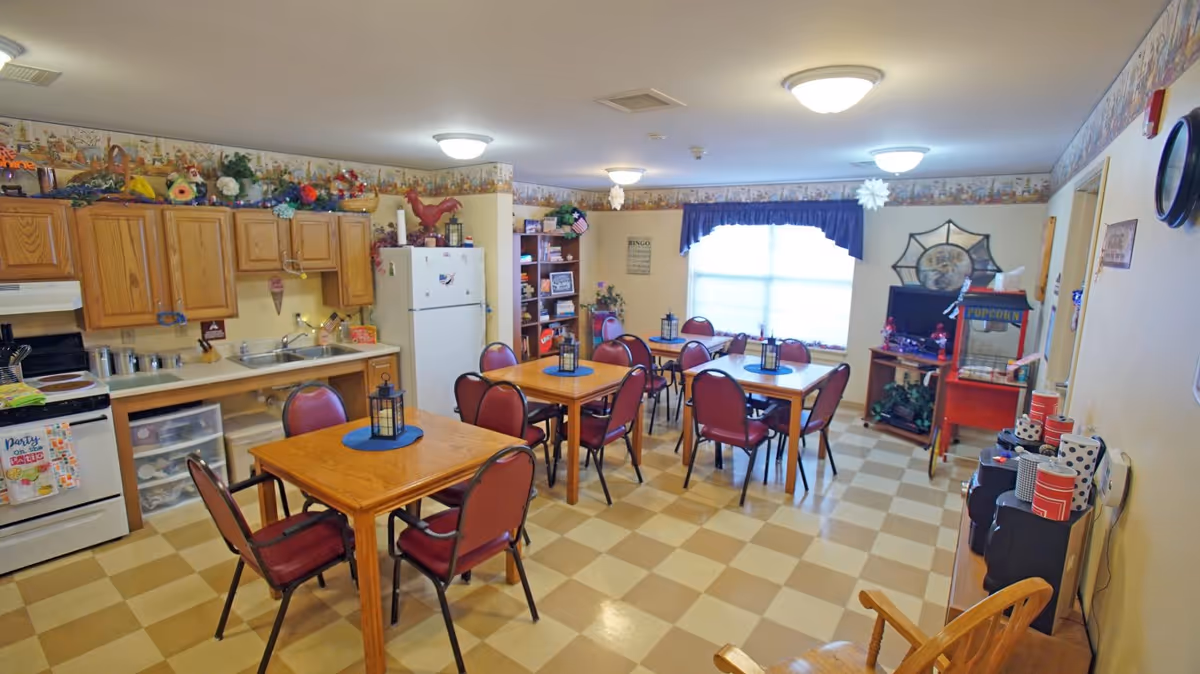 A communal dining area with several wooden tables and red cushioned chairs. The room includes a kitchen area with wooden cabinets, a white refrigerator, and a stove. The floor has a checkered pattern, and the walls are decorated with a wallpaper border near the ceiling. There is a popcorn machine and a TV on a stand near a window with a blue valance. Various decorations and items are placed on shelves and countertops.