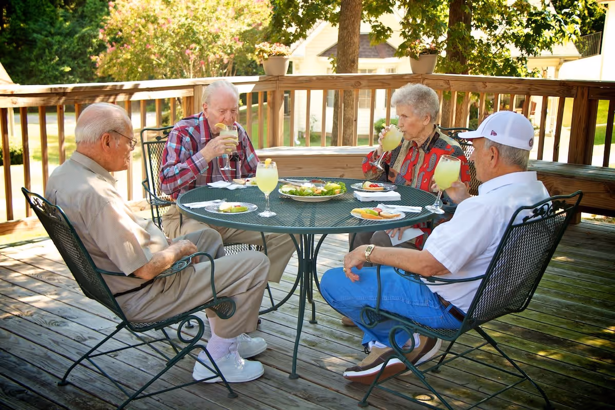 Four elderly adults seated around a metal patio table on a wooden deck, drinking lemonade and sharing a meal.