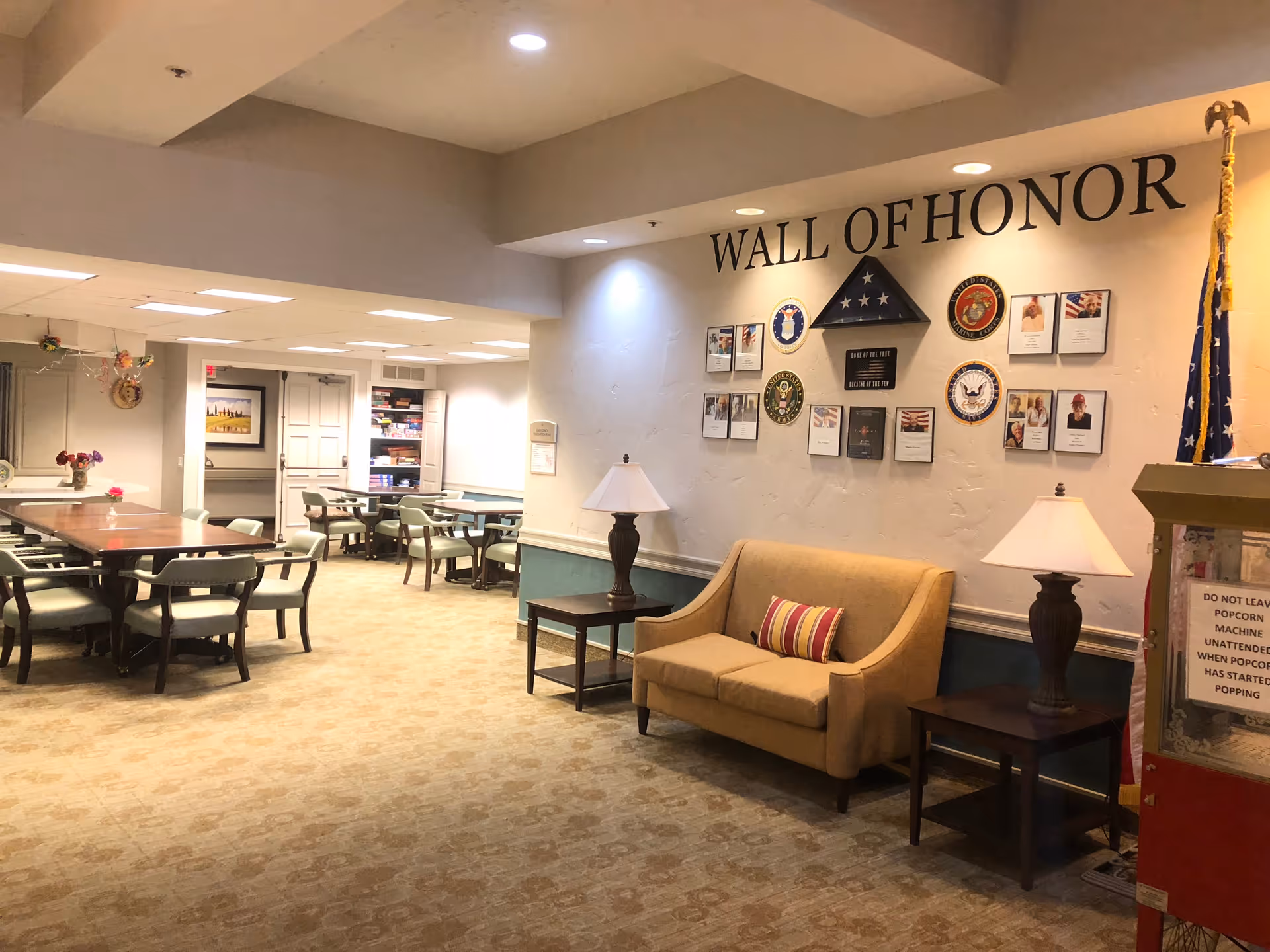 Interior view of a senior living facility common area featuring a 'Wall of Honor' with military insignias and photos, a beige loveseat with a striped pillow, two side tables with lamps, and several tables and chairs in the background for group activities or dining.