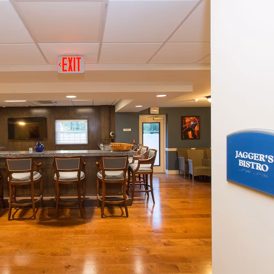 Interior view of Jagger's Bistro at Butler Street Senior Living featuring a bar counter with five wooden bar stools, a TV mounted on a dark wood-paneled wall, a window, and a seating area with chairs and a colorful painting on the wall. An exit sign is visible on the ceiling.