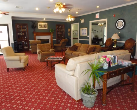 A spacious living room with a red patterned carpet, multiple upholstered sofas and armchairs arranged around a coffee table, a wooden fireplace with built-in bookshelves on either side, ceiling fans with lights, and a side table with a plant and flowers.