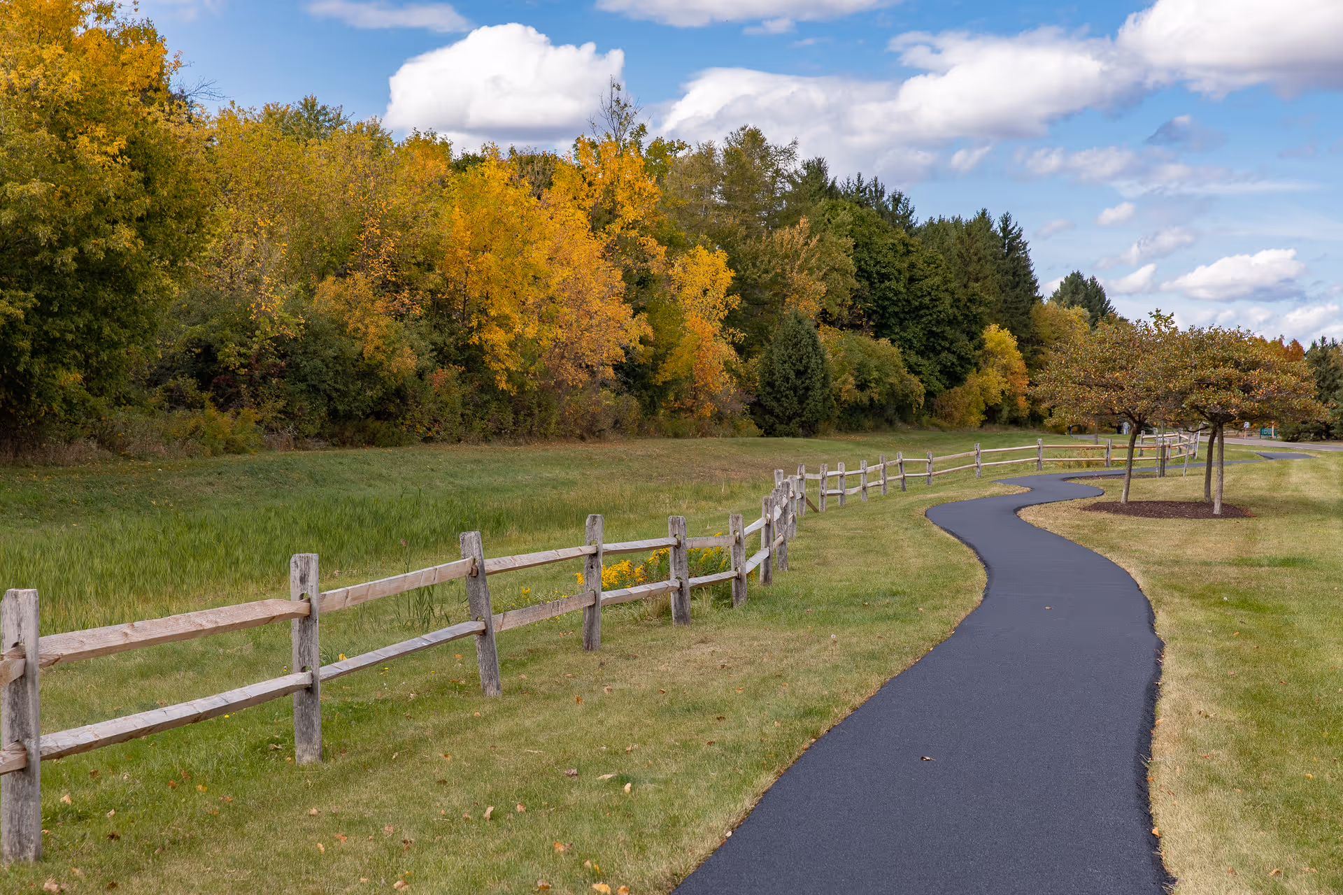 A winding paved pathway bordered by a wooden fence on one side and small trees on the other, with a backdrop of green and yellow autumn trees under a partly cloudy blue sky.