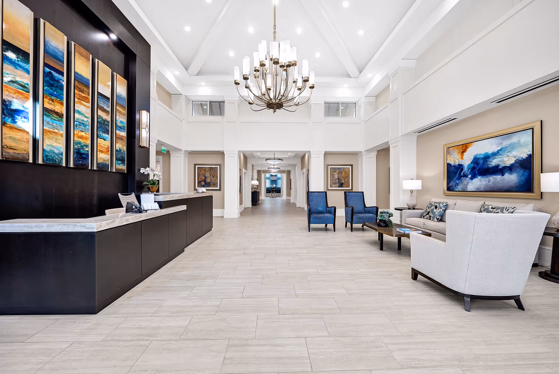 A spacious and bright reception area with a modern chandelier hanging from a high ceiling. The room features a long black reception desk with a marble countertop on the left, colorful abstract artwork on the walls, and a seating area on the right with a sofa, two blue armchairs, a coffee table, and lamps. The floor is tiled in light gray, and the walls are painted in neutral tones with white trim and molding.