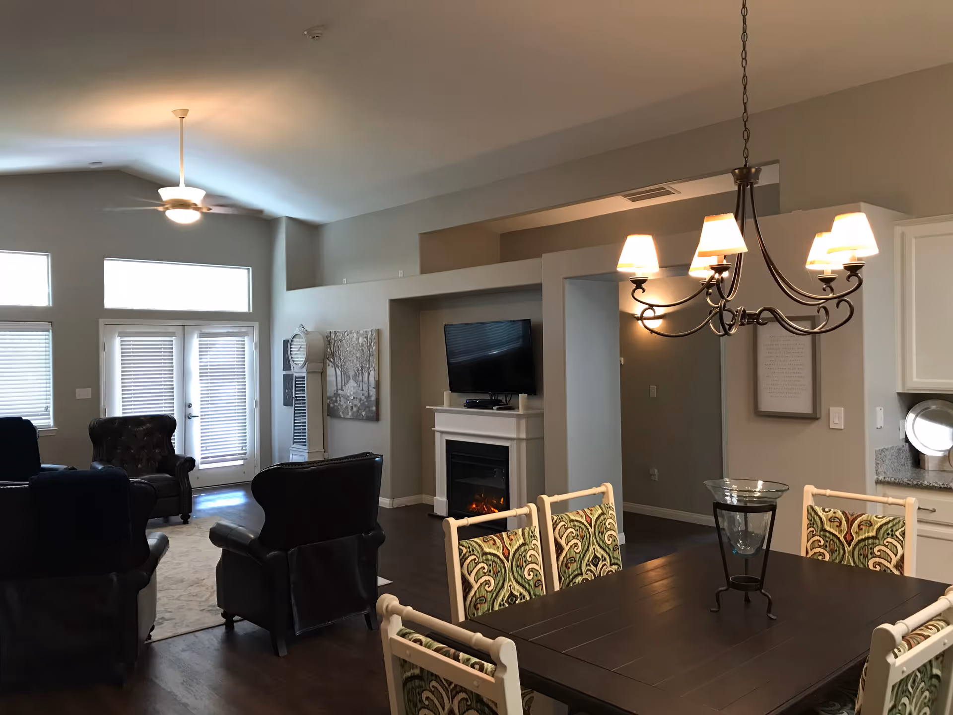 Interior view of a senior living facility showing a combined living and dining area. The living area has several dark leather armchairs arranged around a fireplace with a mounted flat-screen TV above it. The dining area features a dark wooden table with six chairs that have patterned upholstery. A chandelier with five lampshades hangs above the dining table. The room has large windows and glass doors with blinds, light gray walls, and dark wood flooring.