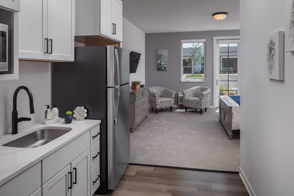 View of a modern senior living apartment featuring a small kitchen area with white cabinets, a black faucet, and a stainless steel refrigerator. Beyond the kitchen is a carpeted living space with two patterned armchairs, a dresser with a small plant and TV mounted on the wall, a window, and a glass door leading outside. Part of a bed is visible on the right side.