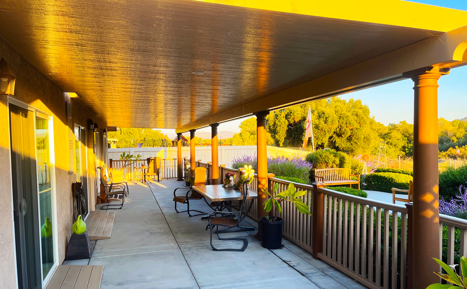 A covered outdoor patio area at a senior living facility with several chairs and tables arranged along the walkway. The patio is supported by brown columns and overlooks a garden with green bushes, purple flowers, and a wooden bench. The scene is bathed in warm sunlight.