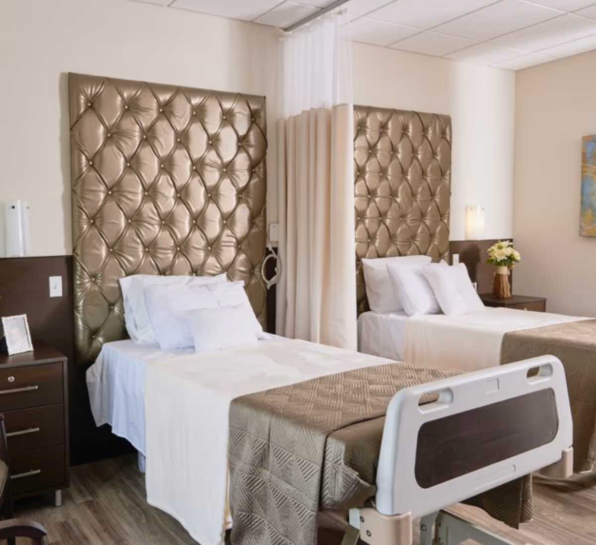 A room with two hospital-style beds separated by a privacy curtain. Each bed has a tall, tufted, metallic gold headboard, white bedding, and a beige quilted blanket. There are nightstands beside each bed, one with a framed photo and the other with a vase of flowers. The room has light-colored walls and wood flooring.