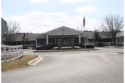 Front exterior view of Lake Park of Madison Nursing and Rehabilitation building with a circular driveway, white fencing, and an American flag on a flagpole in front.