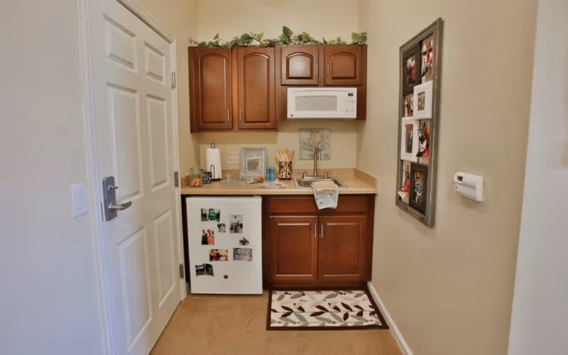 Small kitchenette area with wooden cabinets, a white microwave, a mini refrigerator with photos attached, a countertop with various items including a paper towel holder and a decorative tree picture on the wall, a patterned rug on the floor, and a framed photo collage hanging on the right wall.