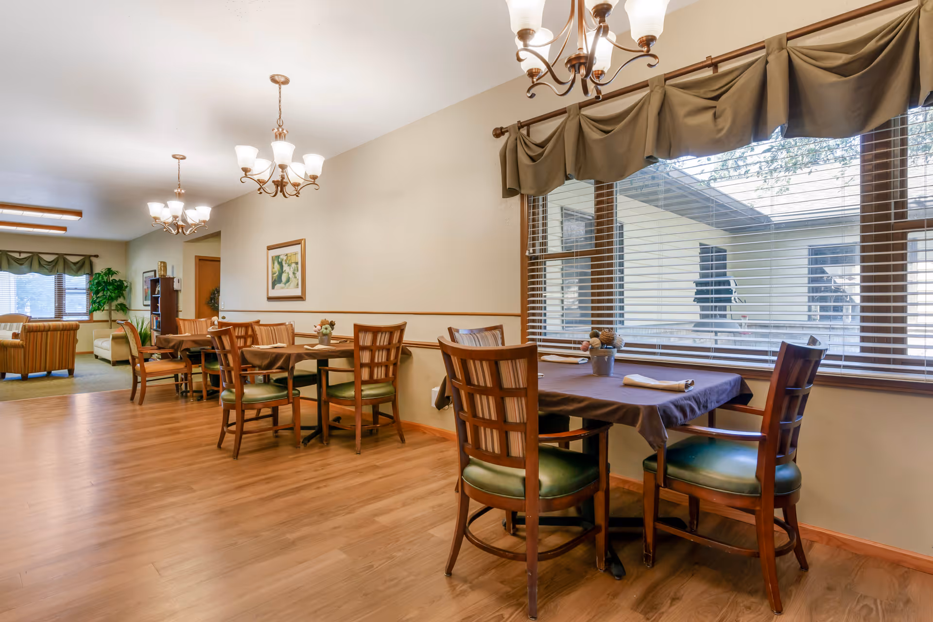 A dining area in a senior living facility with wooden tables and chairs arranged neatly. The tables are covered with dark tablecloths and set with napkins and small flower arrangements. Large windows with blinds and valances allow natural light to fill the room. The space has wooden flooring and warm lighting from ceiling chandeliers. In the background, there is a seating area with armchairs and a bookshelf.