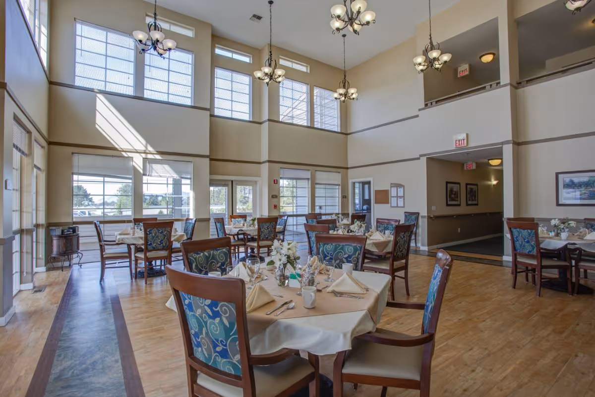 Bright two-story dining room with set tables, patterned chairs, and chandeliers in an assisted living facility.