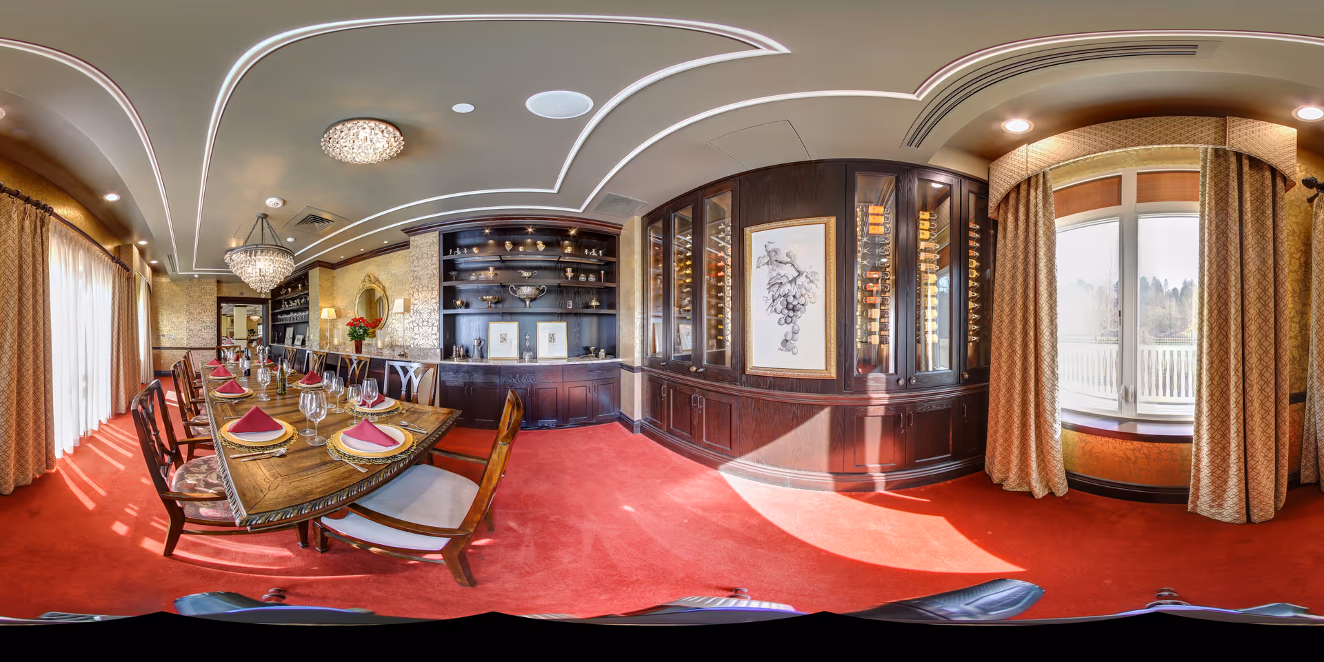 Elegant formal dining room with a long set table, chandeliers, red carpet, built-in dark wood cabinetry and large curtained windows.