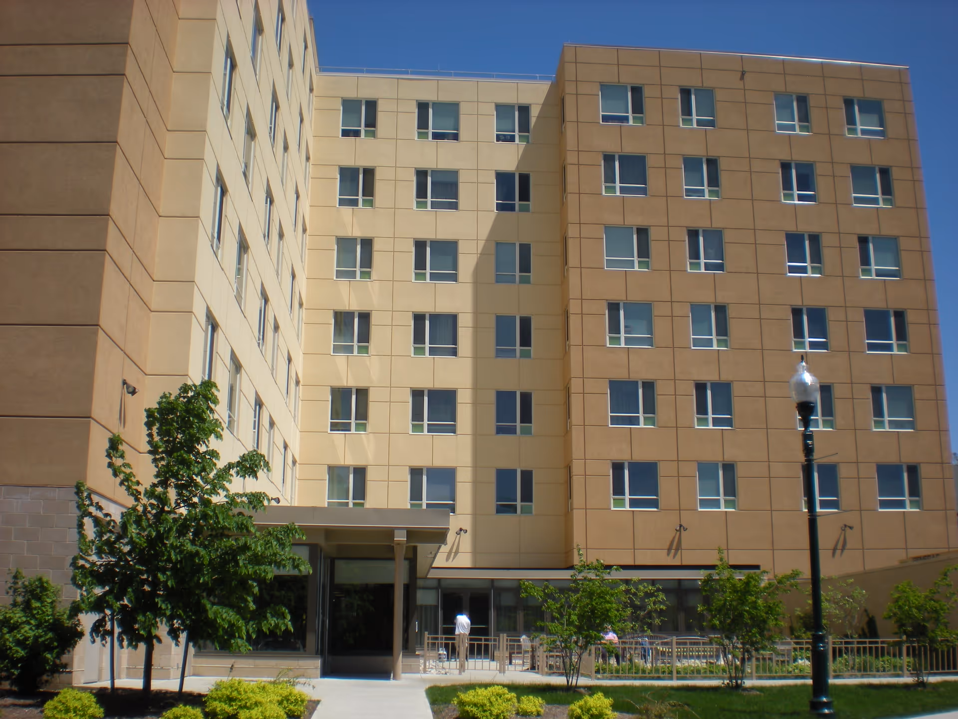 Exterior view of a multi-story senior living facility building with beige and tan walls, multiple windows, a small landscaped area with trees and bushes, a sidewalk leading to the entrance, and a streetlamp in the foreground under a clear blue sky.