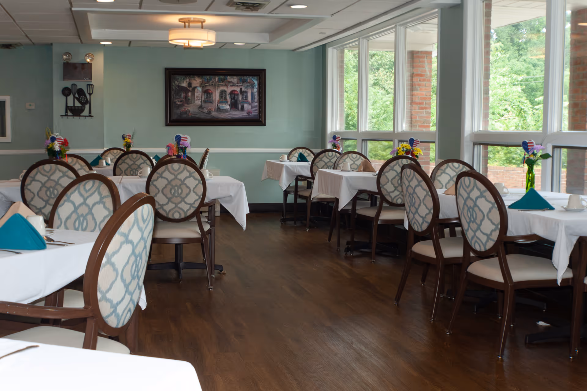 A dining room with multiple tables covered in white tablecloths, each set with cups, napkins folded in a triangular shape, and small flower arrangements with American flag decorations. The room has wooden floors, patterned chairs, large windows letting in natural light, and a painting on the wall.