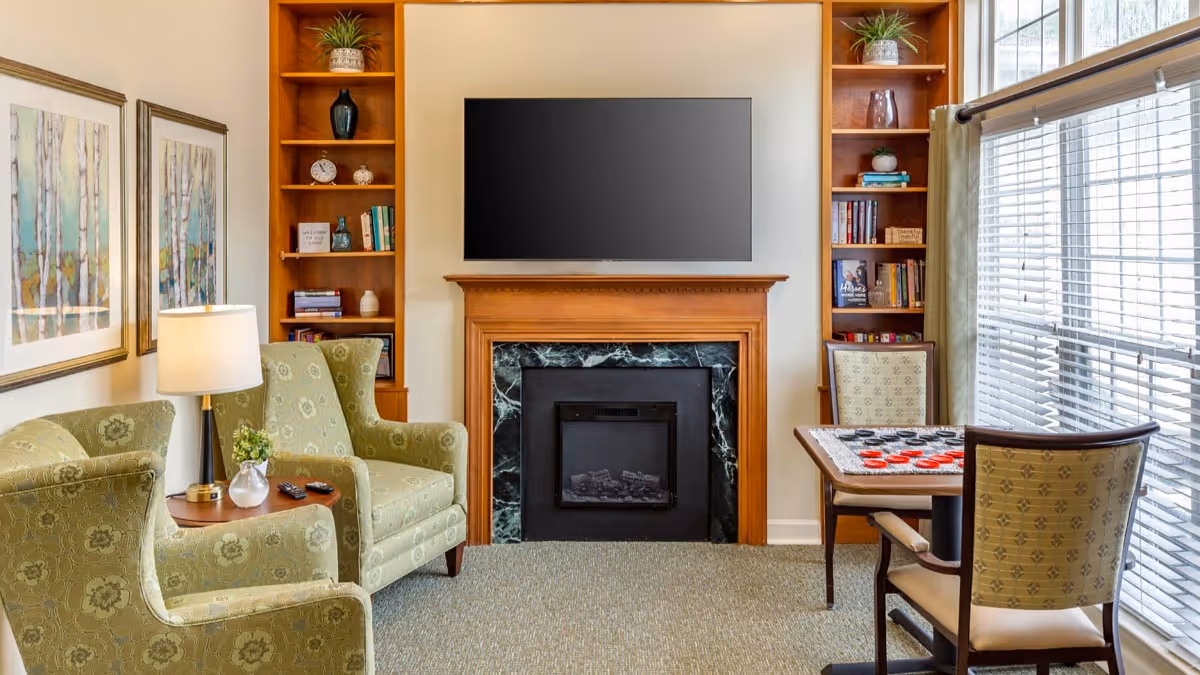 A cozy living room area with two green patterned armchairs and a small wooden side table with a lamp and a small plant. There is a fireplace with a wooden mantel and a black marble surround, above which is a mounted flat-screen TV. On either side of the fireplace are built-in wooden bookshelves with books and decorative items. To the right, there is a small table with a checkers game set up and two chairs, next to a large window with blinds.