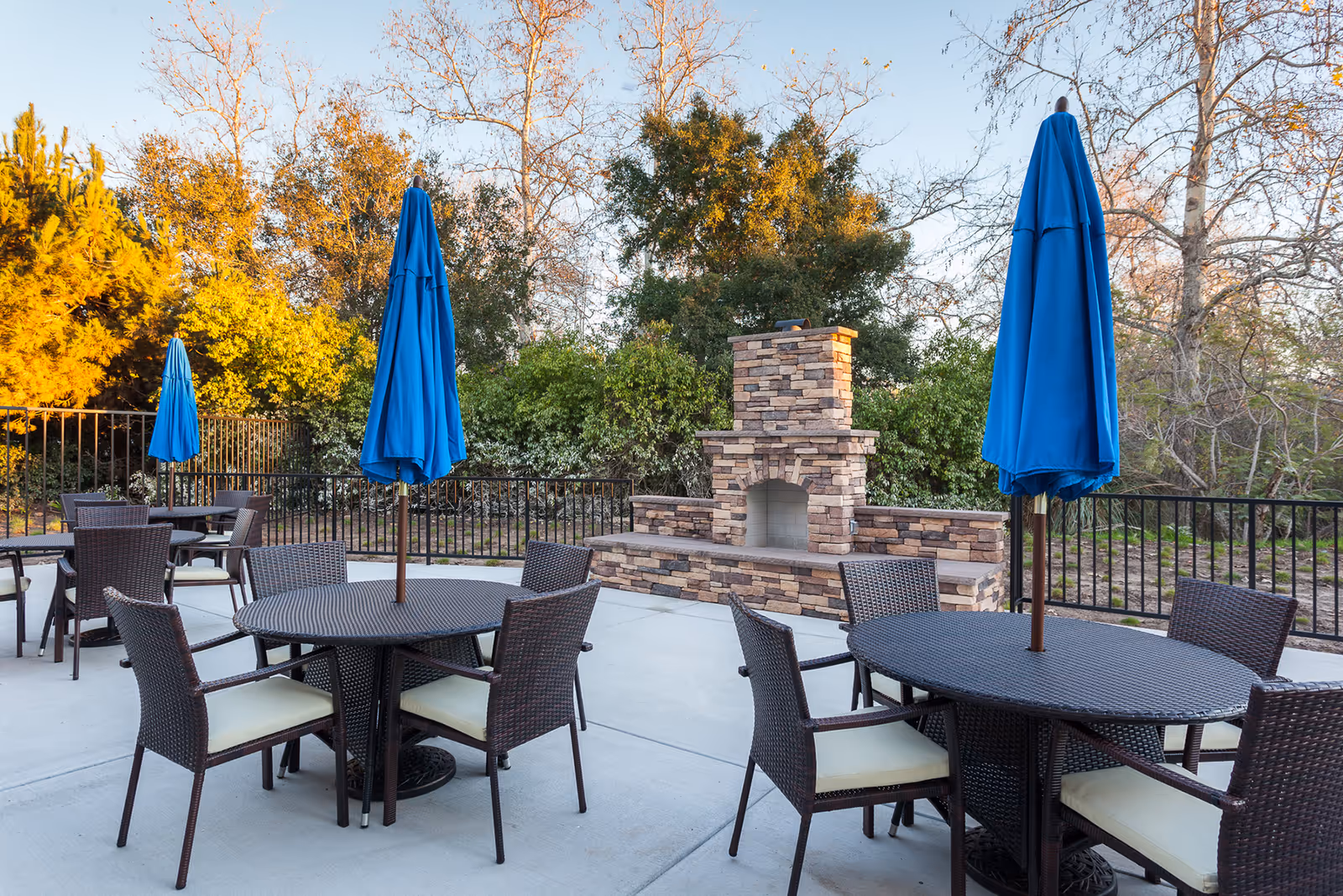 Outdoor patio area with round tables and chairs, each table having a closed blue umbrella. In the background, there is a stone fireplace surrounded by greenery and trees.