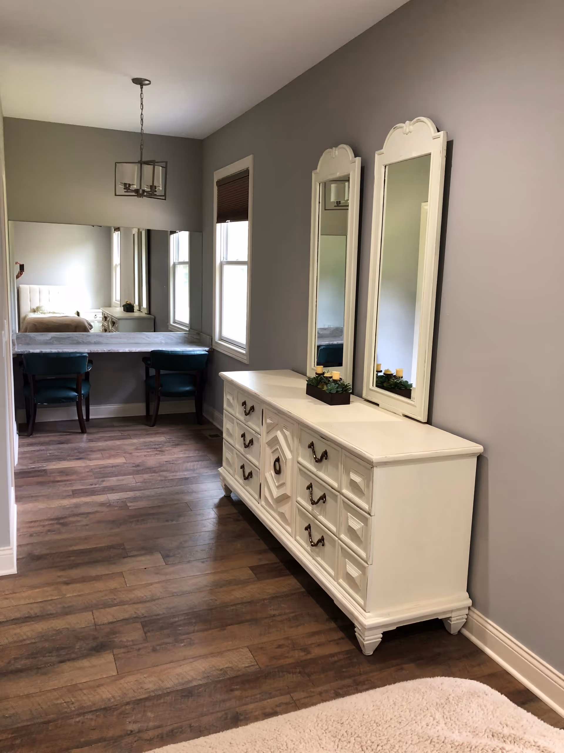 A hallway area in a senior living facility with wooden flooring, light gray walls, and a white dresser with multiple drawers and two tall mirrors above it. In the background, there is a small desk with two teal chairs and a large mirror on the wall reflecting a bed and nightstand with a lamp.