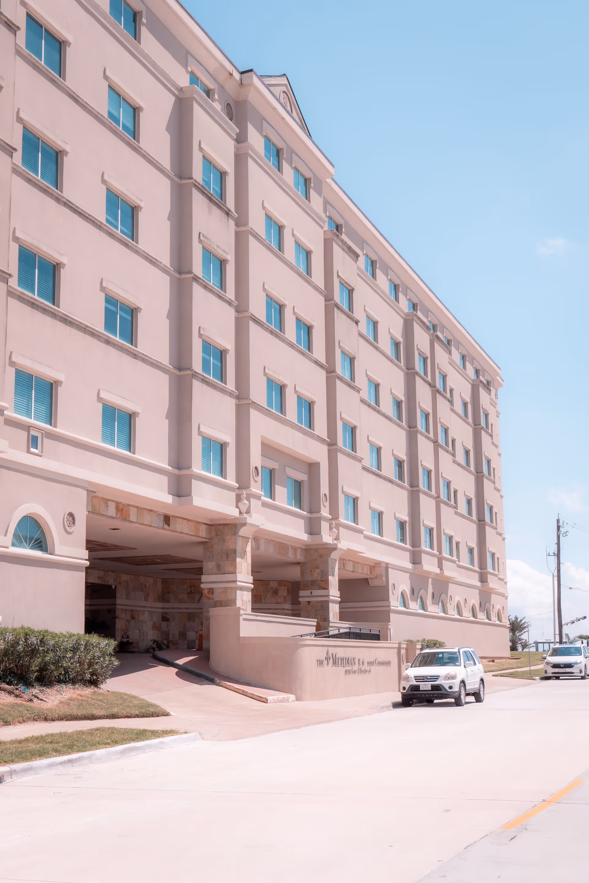 Exterior view of a multi-story beige building named The Meridian with several windows and an entrance driveway. Two white cars are parked on the street in front of the building under a clear blue sky.