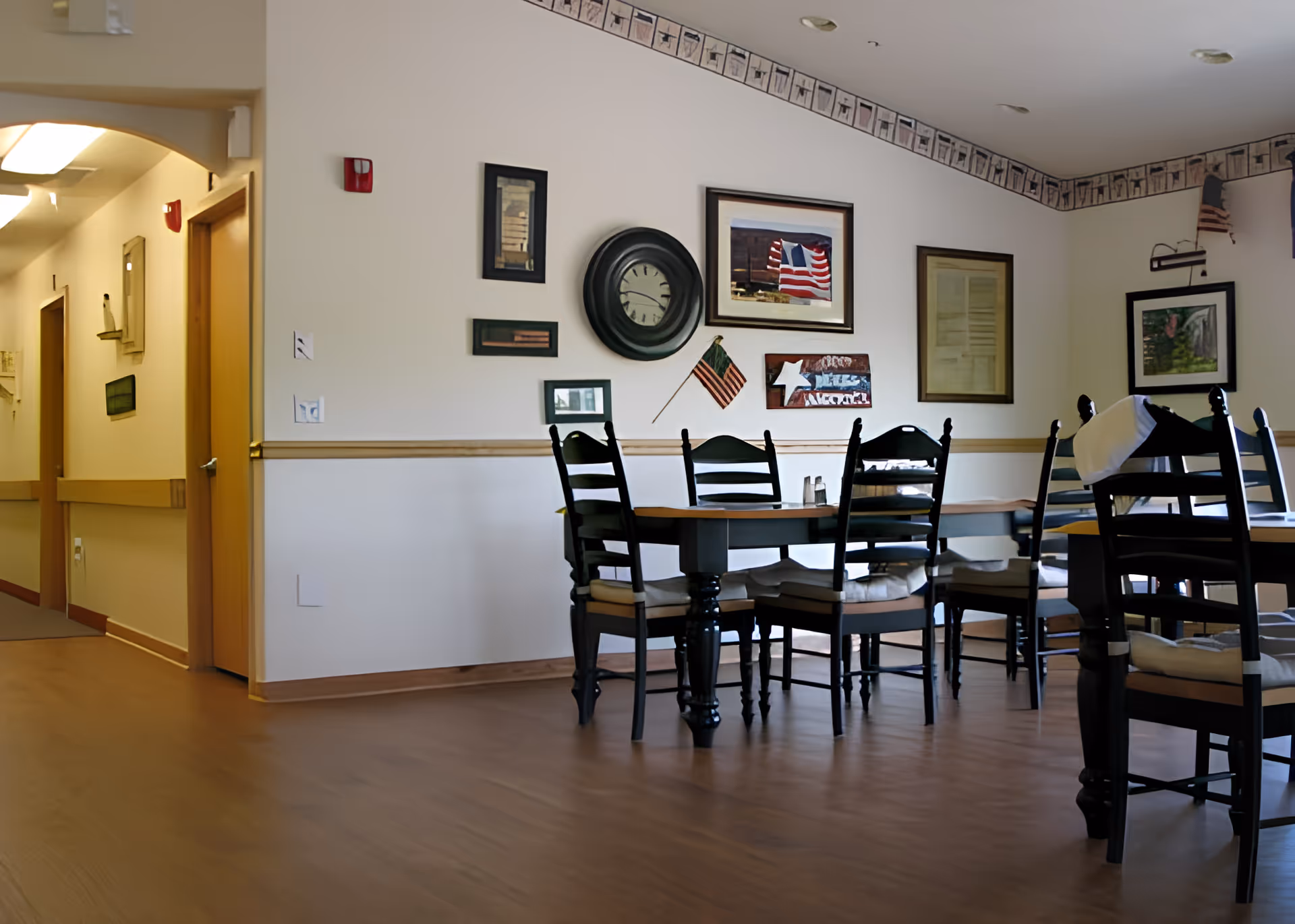 A dining area with a wooden table and several black chairs with cushions. The walls are decorated with framed pictures, a clock, and small American flags. There is a hallway with doors visible to the left.