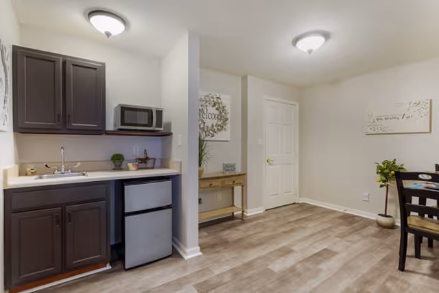 Interior view of a senior living facility kitchen and dining area with dark wood cabinets, a small refrigerator, microwave, sink, and a dining table with chairs. The walls are light-colored with decorative wall art and a small console table with a plant and decor items.