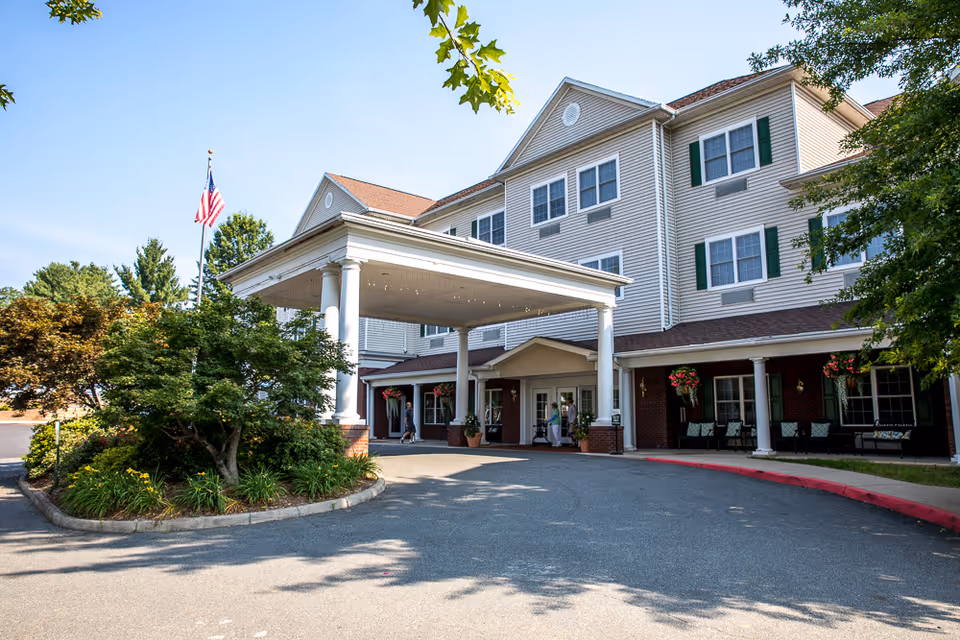 Entrance of a multi-story assisted living facility with a covered drop-off area supported by white columns. The building has beige siding with green shutters and a brick lower facade. There are hanging flower baskets and benches near the entrance, and an American flag is visible on a flagpole to the left. Trees and landscaping surround the driveway.