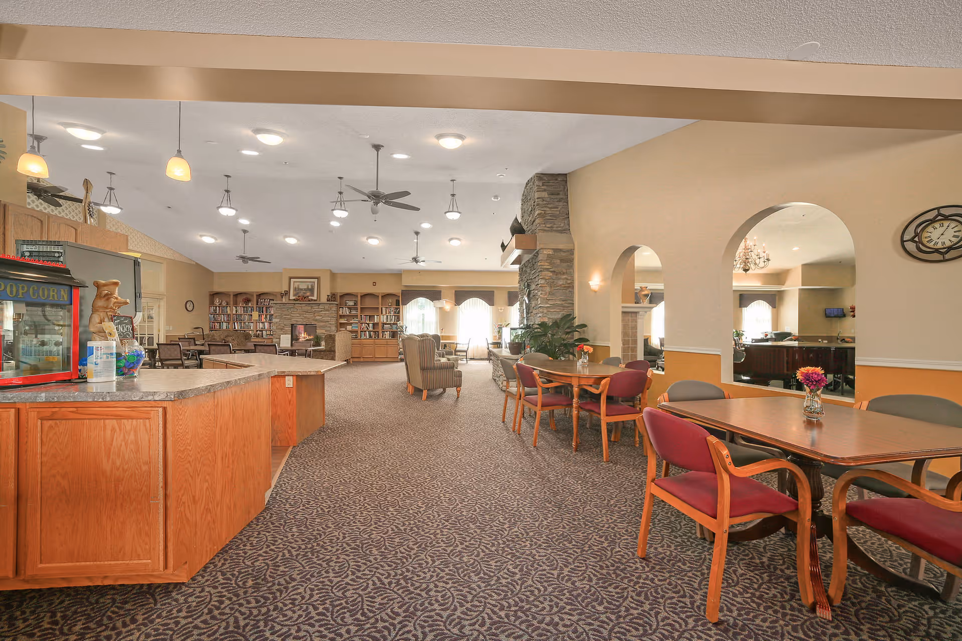 Spacious senior living common area with carpeted floor, multiple tables and chairs, a popcorn machine on a wooden counter, ceiling fans, and a stone fireplace surrounded by bookshelves. The room is well-lit with ceiling lights and natural light coming through large windows.