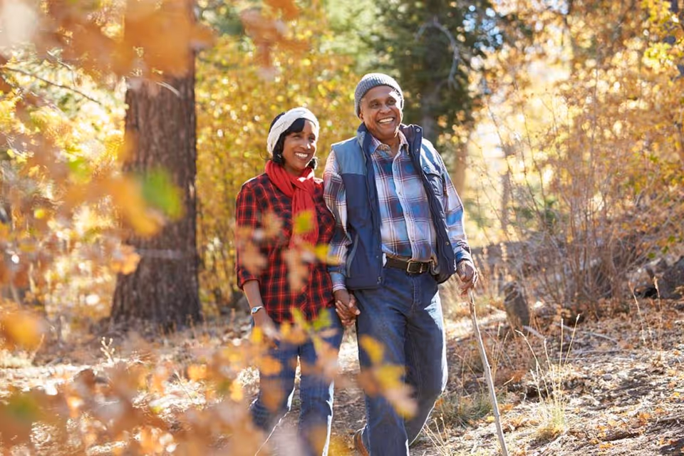 An elderly couple holding hands and walking outdoors through a wooded area with autumn foliage. The man is wearing a gray beanie, blue vest, and plaid shirt, and the woman is wearing a white knit hat, red scarf, and red plaid shirt. They are smiling and enjoying the walk together.