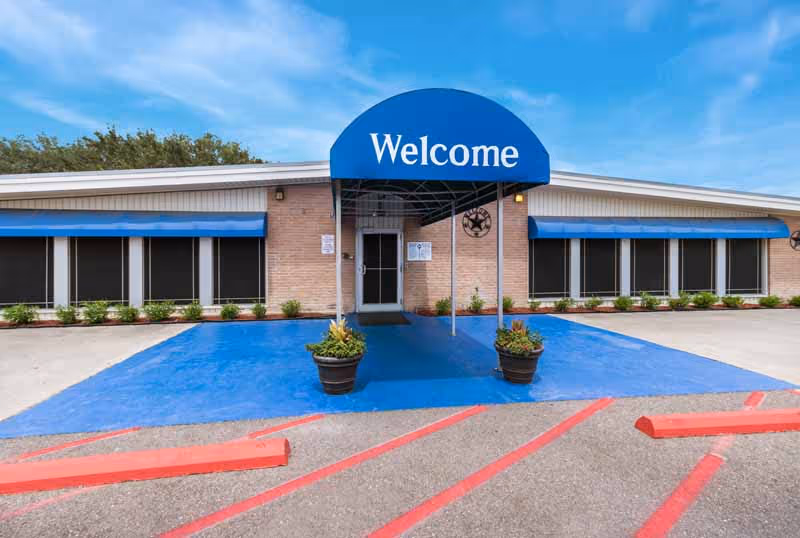 Front entrance of a single-story nursing facility with a blue 'Welcome' awning over the doorway and planters on a painted blue entryway.