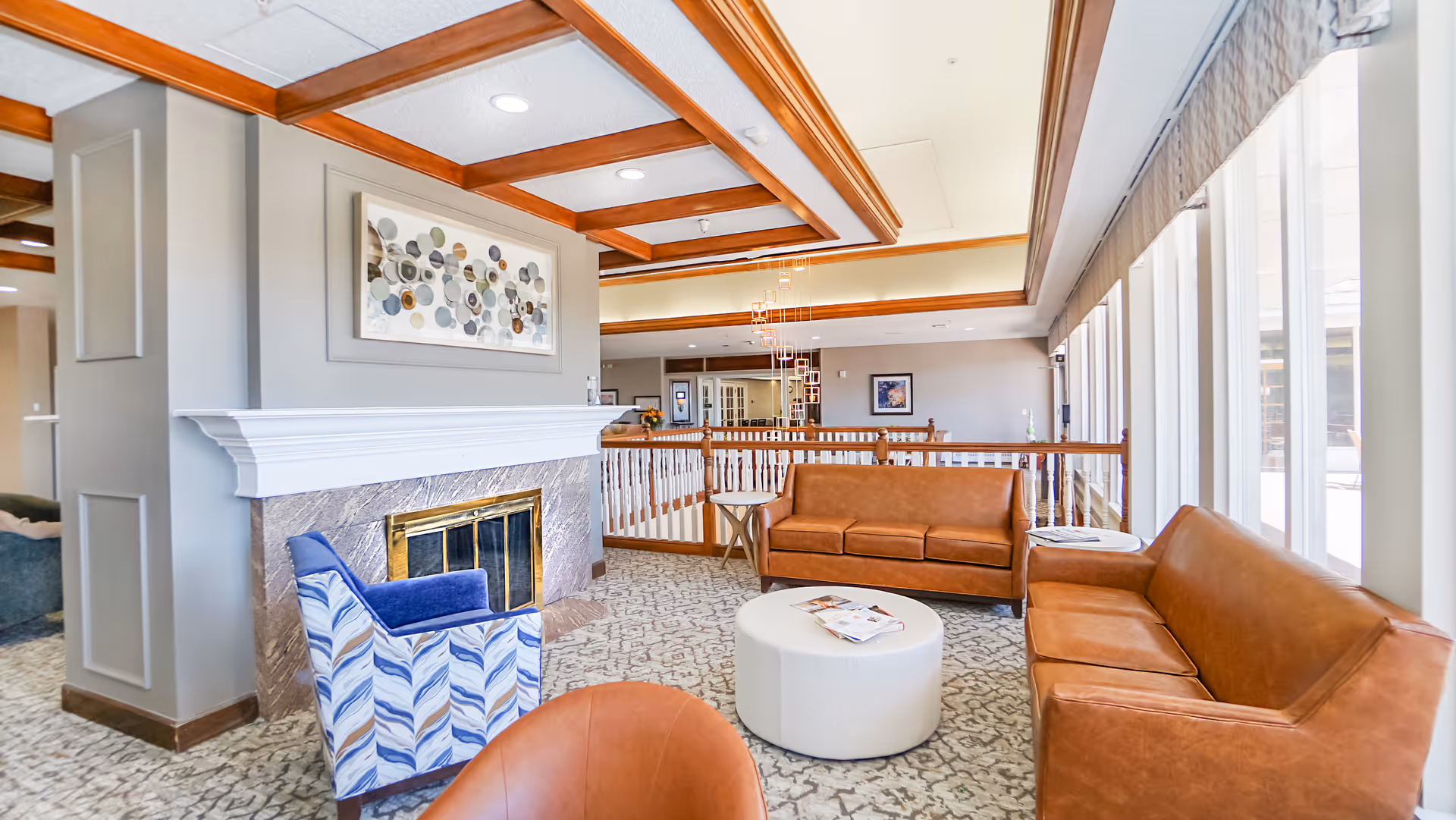 Sunlit communal lounge with leather sofas, a patterned armchair, fireplace, and a round coffee table by large windows.