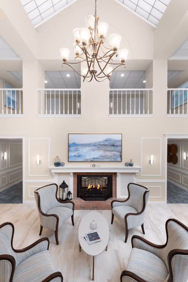 A bright and elegant living room area with four upholstered armchairs arranged around a small oval coffee table. The room features a lit fireplace with a white mantel, decorated with small vases and a framed landscape painting above it. There is a large chandelier hanging from the high ceiling with skylights, and a second-floor balcony with white railings overlooking the space.