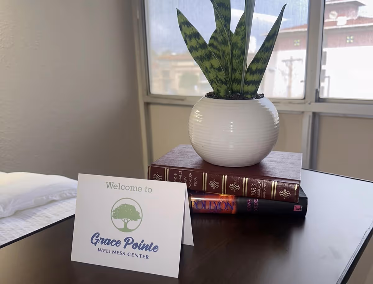 A small table by a window holds a potted plant on stacked books and a card that reads 'Welcome to Grace Pointe Wellness Center' next to a bed.