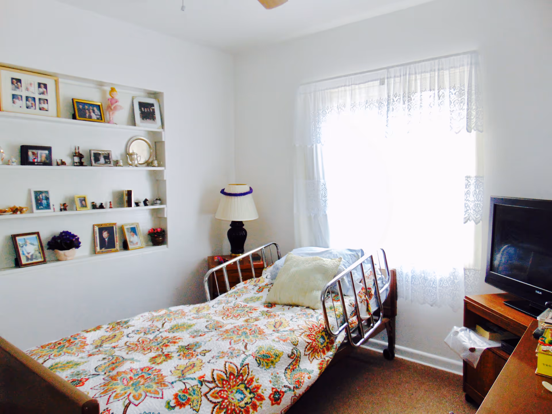A small bedroom with a single bed covered in a colorful floral bedspread. The bed has metal rails and is positioned near a window with white lace curtains. There is a wooden nightstand with a lamp and a wooden dresser with a television on top. Built-in shelves on the wall display framed photos, small figurines, and decorative items.