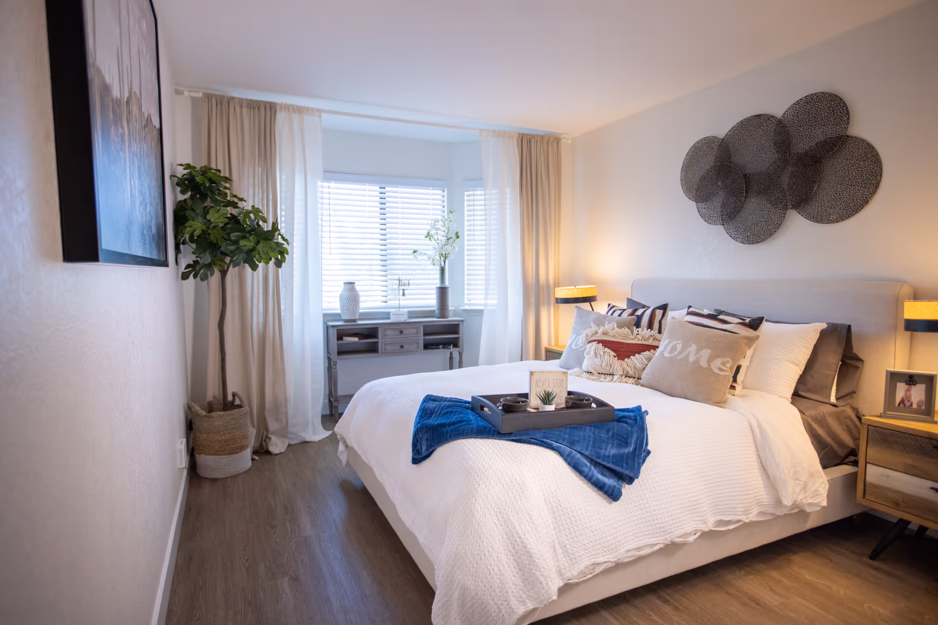 A bright and cozy bedroom with a large bed covered in white bedding and multiple decorative pillows. A blue throw blanket and a tray with small items are placed on the bed. The room features wooden flooring, a potted plant in the corner, a small desk with decorative vases near a window with white blinds and curtains, and two bedside tables with lamps. A modern circular wall art piece is mounted above the bed.