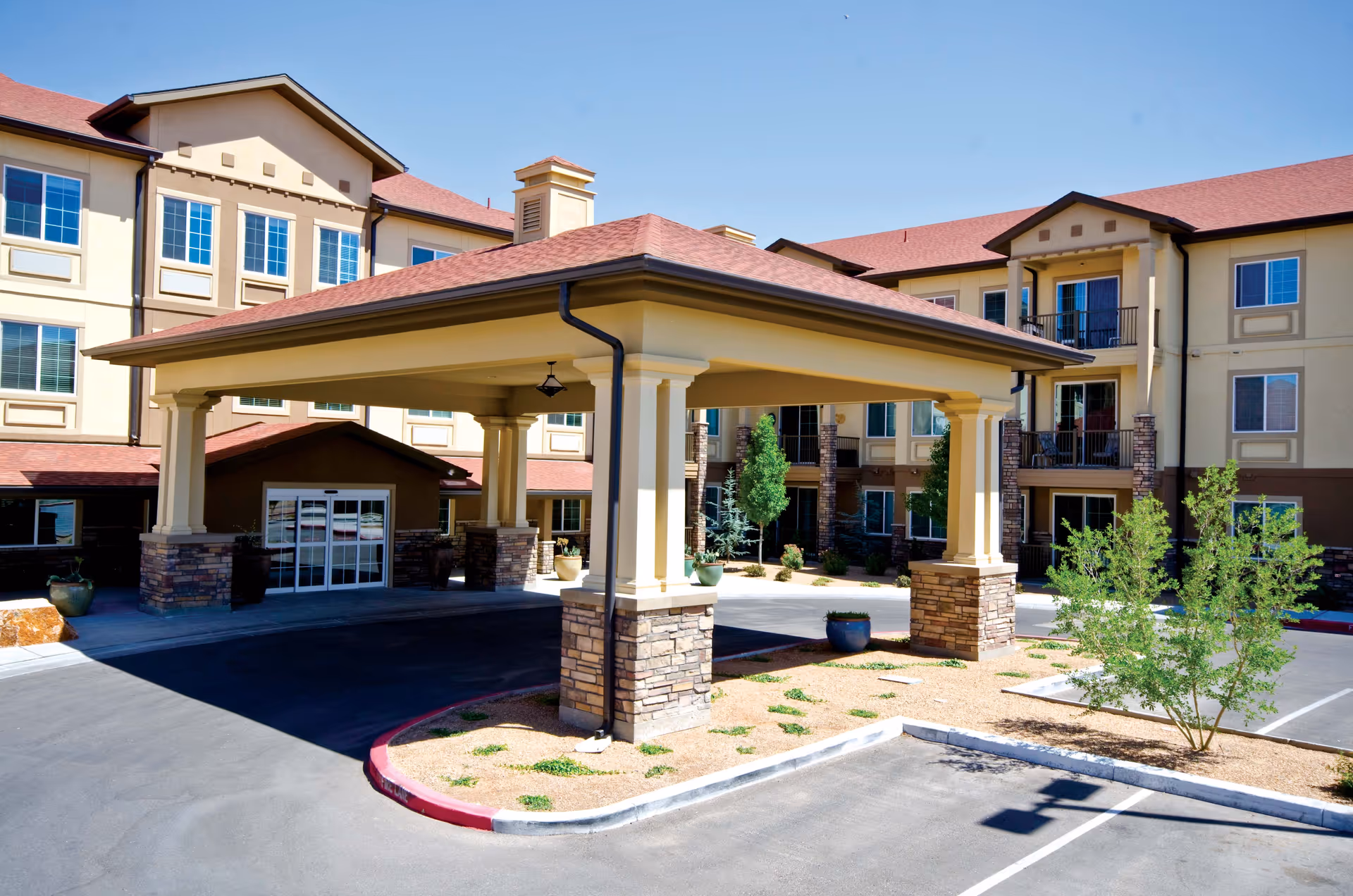 Exterior view of Palmilla Senior Living facility showing a covered entrance with stone pillars, multiple windows, balconies, and a parking area with some landscaping including small trees and plants under a clear blue sky.