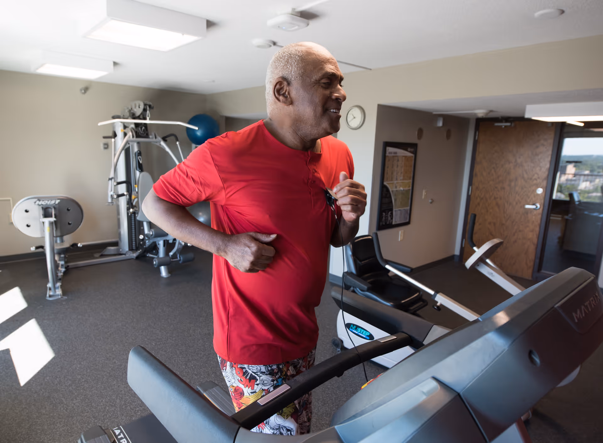 An elderly man wearing a red shirt and patterned shorts is jogging on a treadmill in a fitness room equipped with various exercise machines and equipment. The room has beige walls, a clock on the wall, and a wooden door with a window showing an outside view.