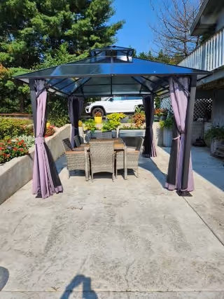 Outdoor patio area with a metal-framed gazebo featuring a transparent roof and purple curtains tied back at the corners. Underneath the gazebo is a rectangular table surrounded by six wicker chairs. The patio is surrounded by greenery and flowers, with a white vehicle and part of a building visible in the background.