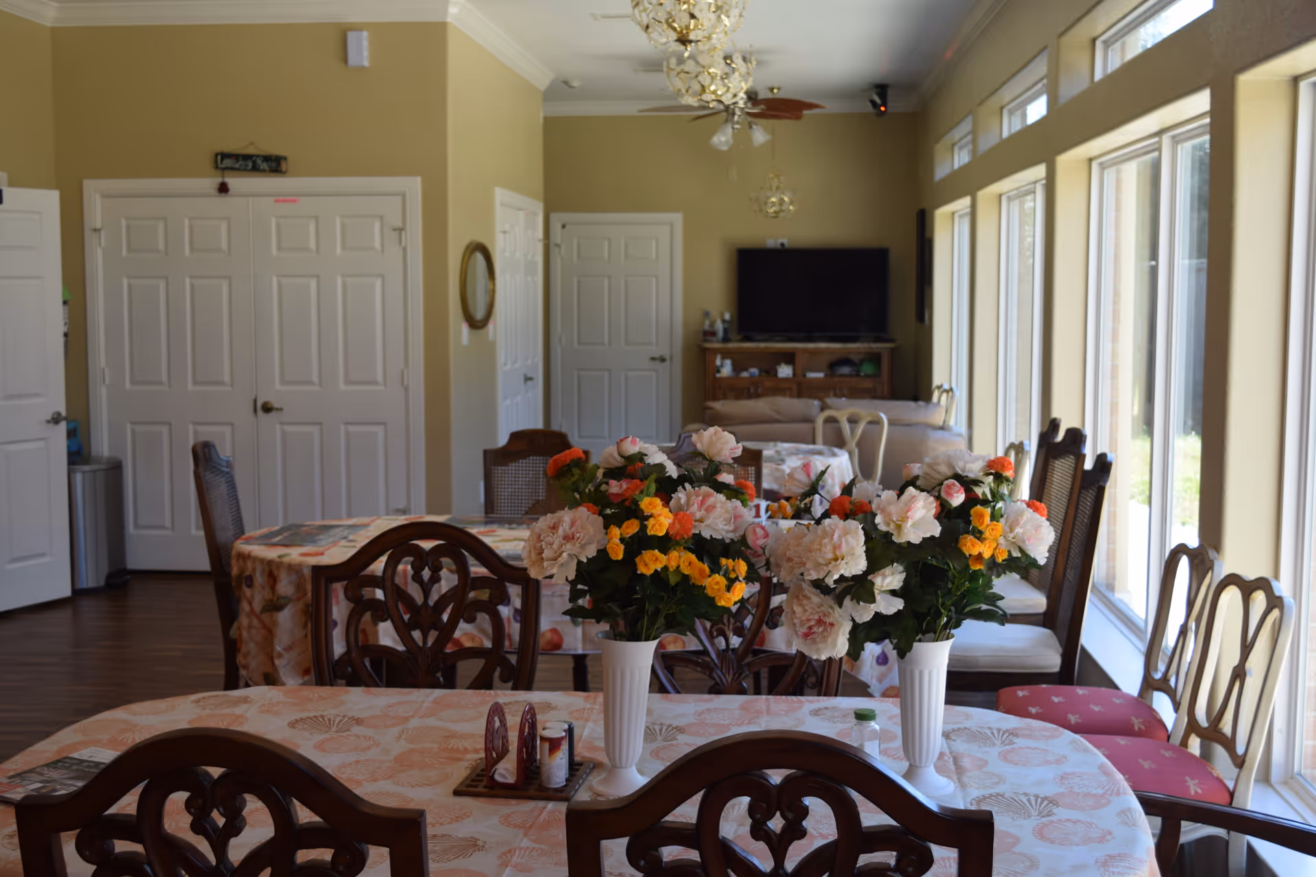 A bright dining area in an assisted living facility with multiple tables covered in patterned tablecloths. Each table has vases filled with colorful artificial flowers. The room has large windows on one side allowing natural light to fill the space. In the background, there is a living area with a TV and beige couches. The walls are painted light yellow, and there are ceiling fans with lights hanging from the ceiling.