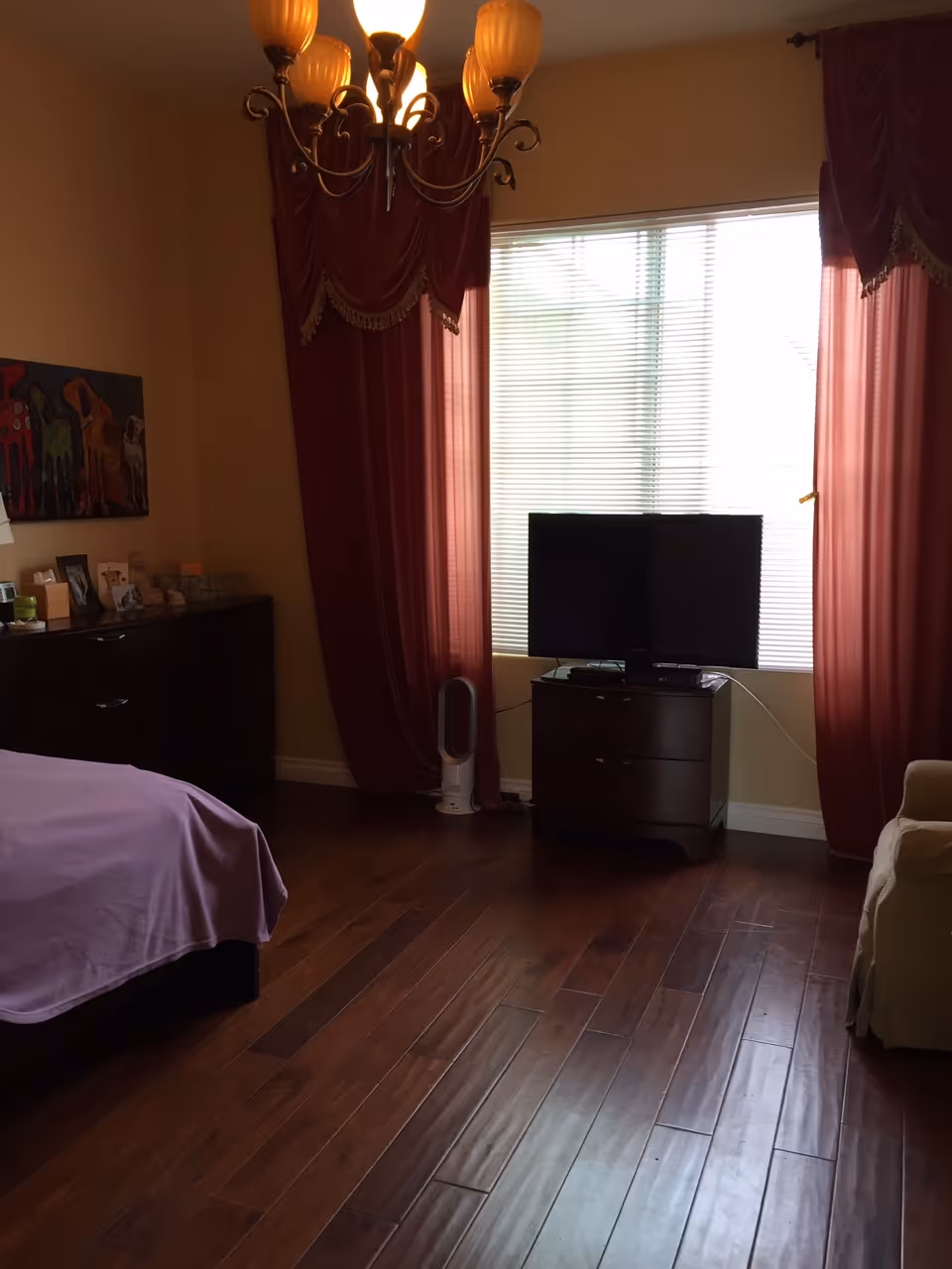 A bedroom with wooden flooring, a bed covered with a purple blanket on the left, a dark wooden dresser with framed photos and decorations, a TV on a small dark wooden stand in front of a window with closed blinds and red curtains, and a beige armchair on the right. A chandelier with multiple lights hangs from the ceiling.