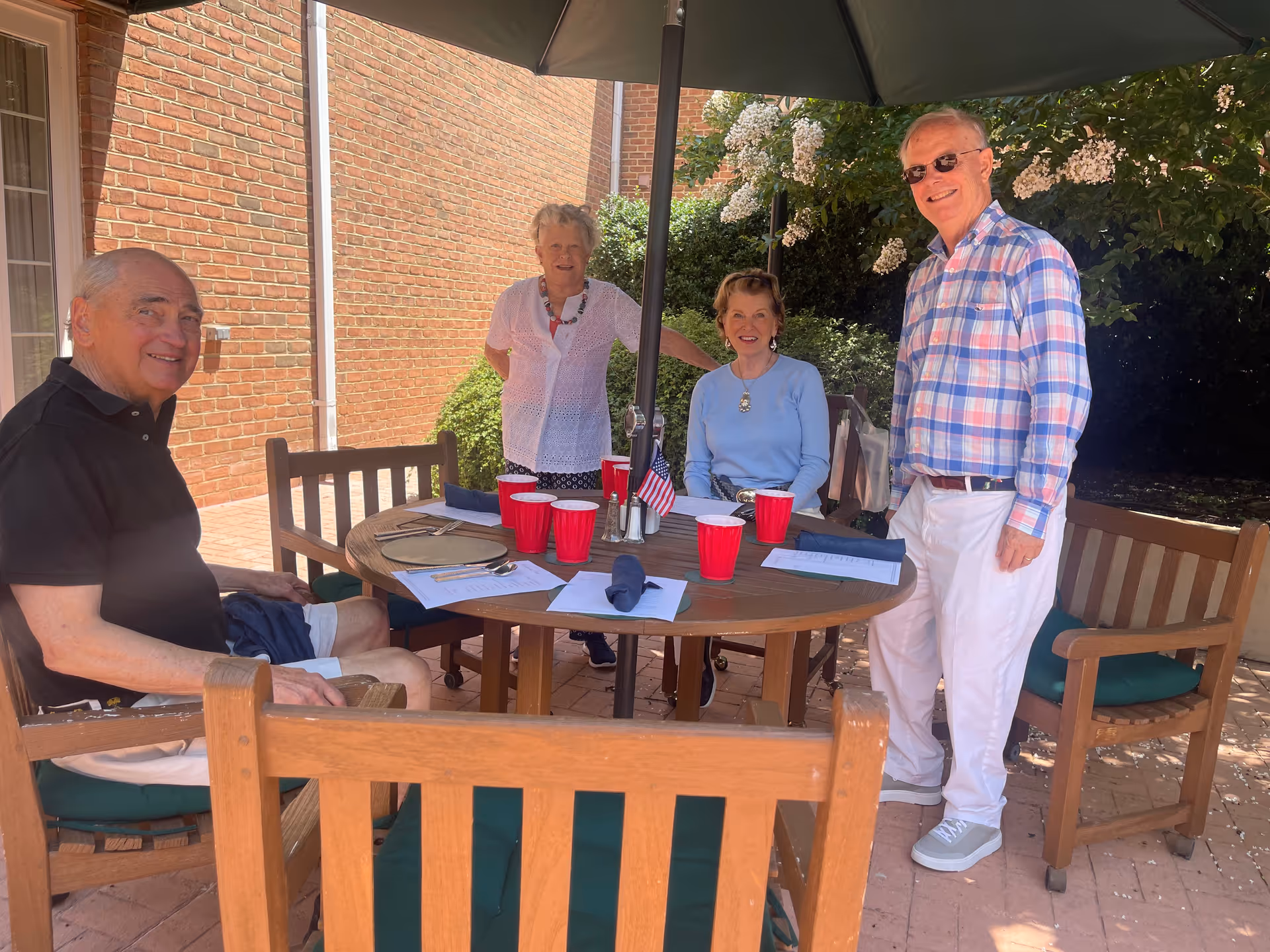Four elderly people gathered around a wooden outdoor table with an umbrella. The table has red plastic cups, napkins, and papers on it. They are smiling and appear to be enjoying a sunny day in a patio area with brick walls and greenery in the background.
