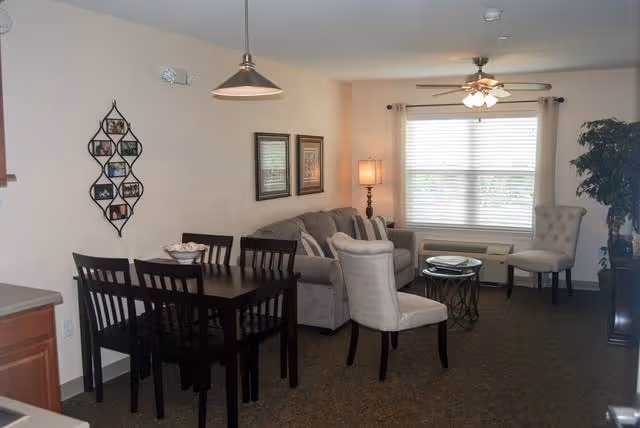 Well-lit combined living and dining area with a dark wood dining table, sofa and accent chairs, coffee table, and a window with blinds.