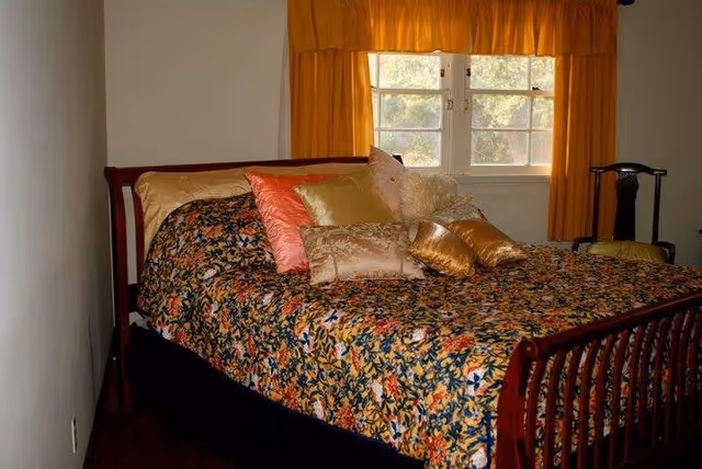 A bedroom with a wooden bed frame featuring a floral patterned bedspread in dark colors with multiple decorative pillows in gold, pink, and beige. Behind the bed is a window with yellow curtains, and to the right side of the bed is a wooden chair with a yellow cushion.