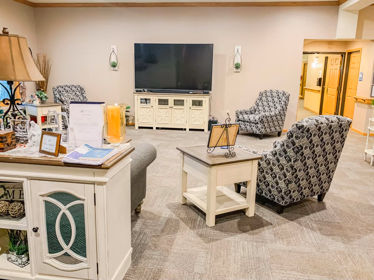 A cozy living room area in Meadow View Assisted Living featuring a large flat-screen TV mounted on a cream-colored cabinet. The room has patterned armchairs, a small wooden side table with a decorative frame, and a white cabinet with various decorative items including a lamp and a large candle. The walls are beige and the carpet is light brown with a subtle pattern. The space is well-lit and has an inviting atmosphere.