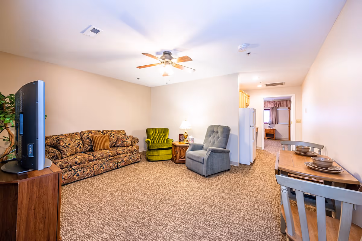 A cozy living area in an assisted living facility featuring a floral patterned sofa, a green armchair, a gray recliner, a wooden side table with a lamp, a flat-screen TV on a wooden stand, and a small dining table with two chairs set with bowls and plates. The room has beige walls, carpeted flooring, and a ceiling fan with lights. A kitchen area with a refrigerator is visible in the background.